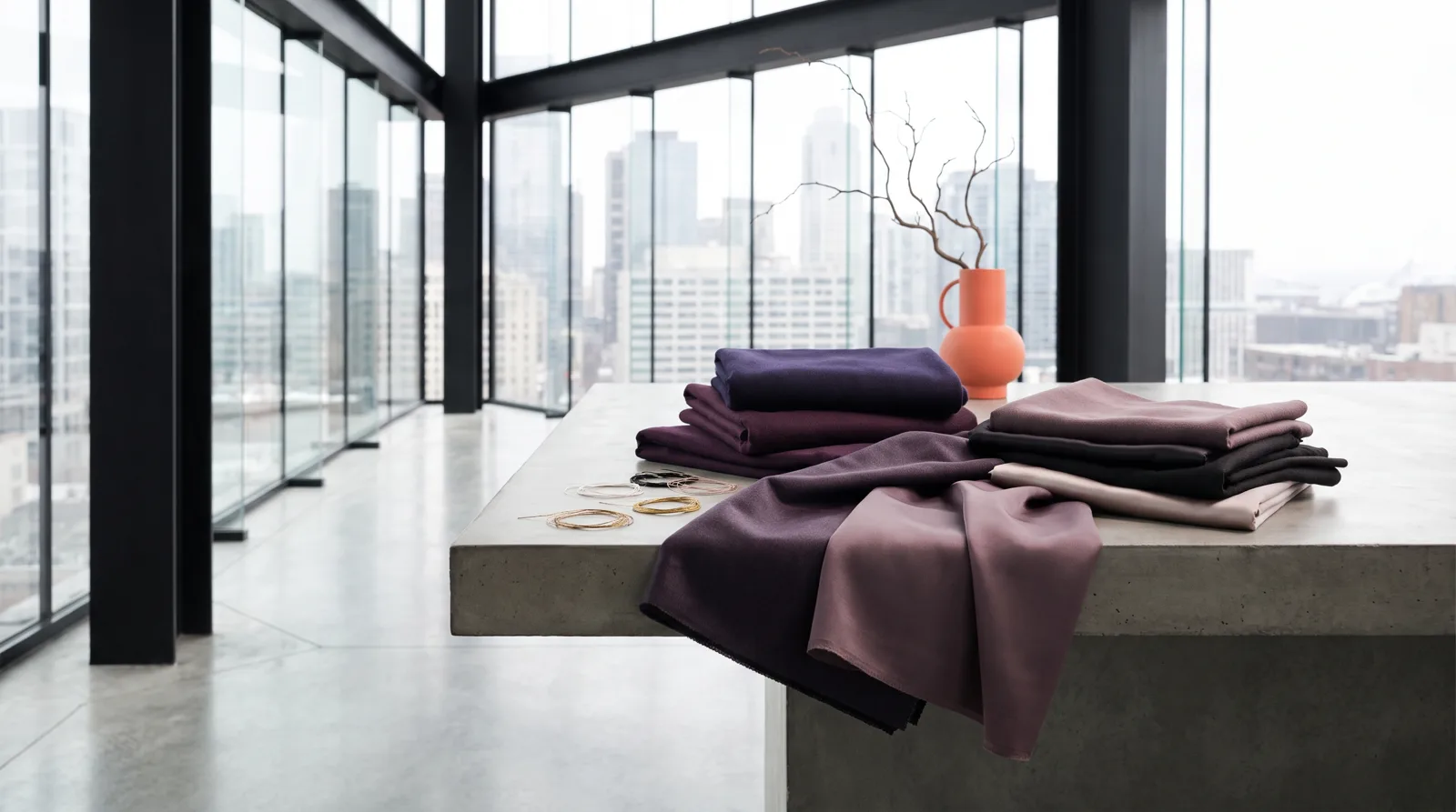 Gefaltete Stoffmuster in tiefem Violett, Pflaume und Mauve auf einem Betontisch, dazu goldene Metallfäden
